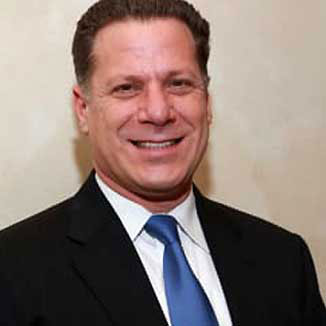 A man wearing a suit and tie stands smiling against a beige background with a blue ribbon.