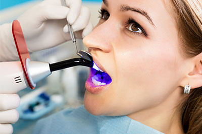 The image shows a person seated with an open mouth under a dental device, being attended by a professional wearing medical gloves and using a device to examine or treat the individual s teeth.