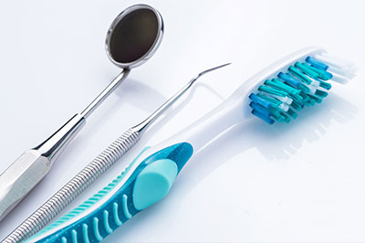 The image displays a collection of dental instruments including a toothbrush with blue bristles, dental picks, a dental mirror, and a pair of tweezers, all arranged on a white surface against a light background.