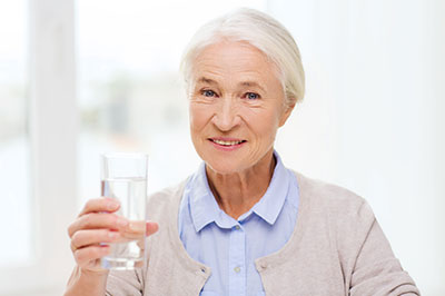 The image features an elderly woman holding a glass of water with both hands, smiling at the camera.