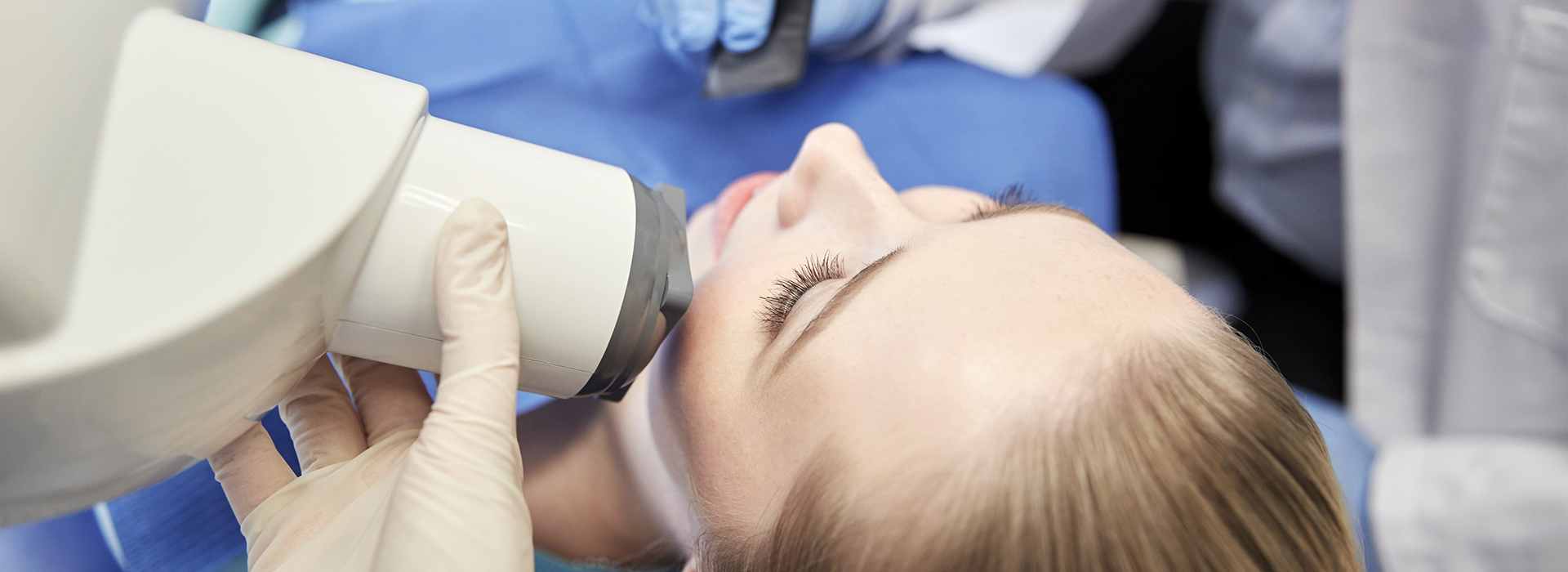 A woman is sitting in a dental chair with her eyes closed while a dentist performs a procedure on her teeth using a magnifying device.