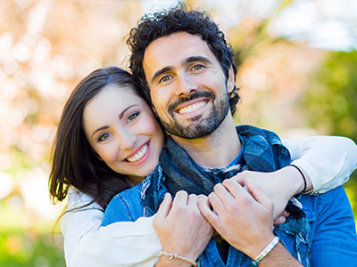 A man and woman are hugging each other warmly outdoors, with the man wearing a bandana around his neck.