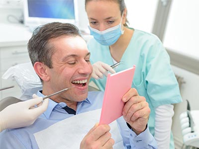 A man sitting in a dental chair with a smiling expression, holding up a pink card, while a woman in scrubs looks on.