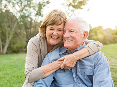 The image depicts an older man and woman embracing each other with affectionate smiles, set against a backdrop of a serene outdoor setting during daylight hours.