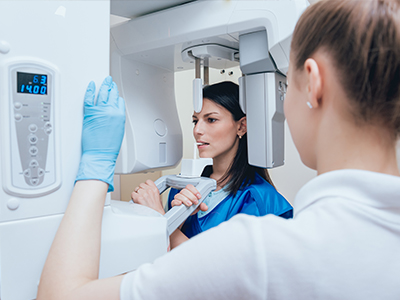 A woman in blue scrubs stands beside a large, modern 3D scanner, with a person wearing a white lab coat observing from behind.