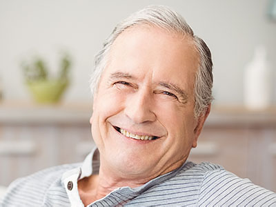 The image shows a smiling older man with gray hair, wearing a blue shirt, sitting comfortably in a chair indoors, with a relaxed posture and looking directly at the camera.