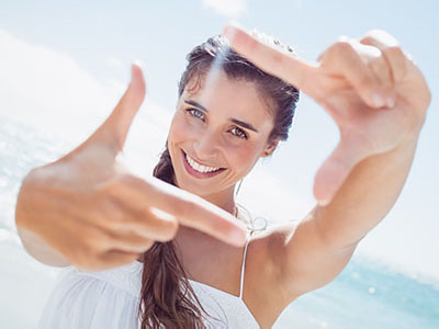 A woman with long hair is smiling at the camera while holding up her hand to frame a picture of herself against a sunny beach backdrop.